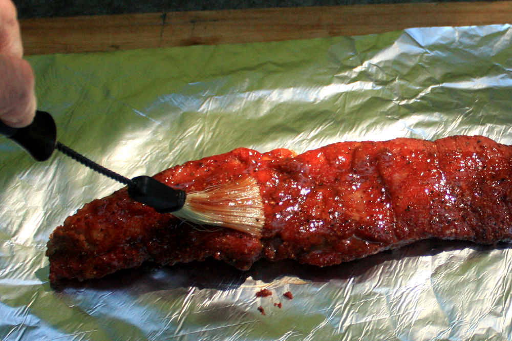 Brushing partially cooked smoked pork brisket with bbq sauce before foil-wrapping to finish cooking in the smoker.